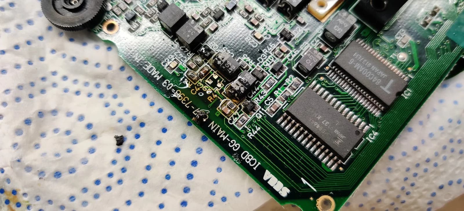 A cleaned-up area of a Game Gear board where flux was used to lift electrolyte contamination from the SMD solder joints, the transistor Q9 missing from its footprint after the contamination there loosened it during cleaning, its blackened legs visible on the mat beside the board.