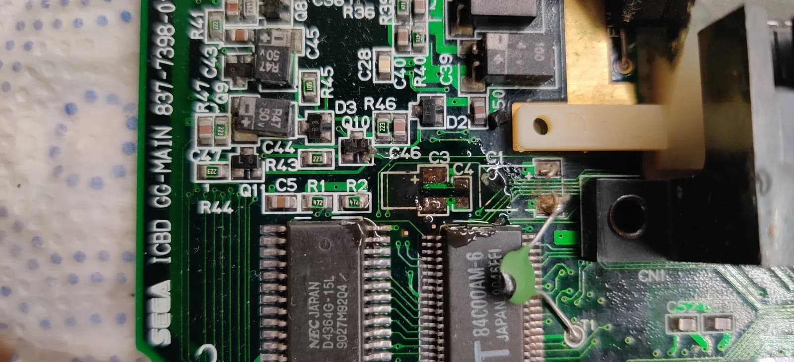 A Game Gear board during cleanup with the capacitors C3 and C1 just removed, their pads heavily contaminated, and the surrounding SMD parts and electrolytic capacitors all showing corroded, contaminated solder joints.
