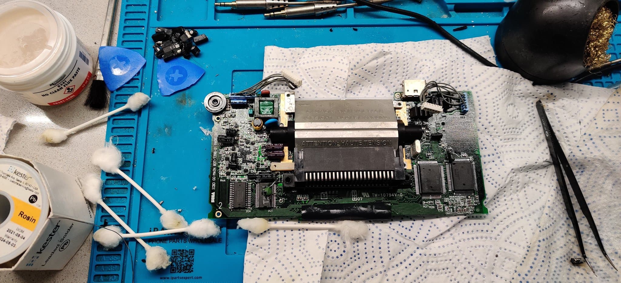 A single Game Gear board, now fully recapped, with the old capacitors piled on the blue mat at top left and used cotton swabs from fluxing and cleaning the corroded solder joints beside it.