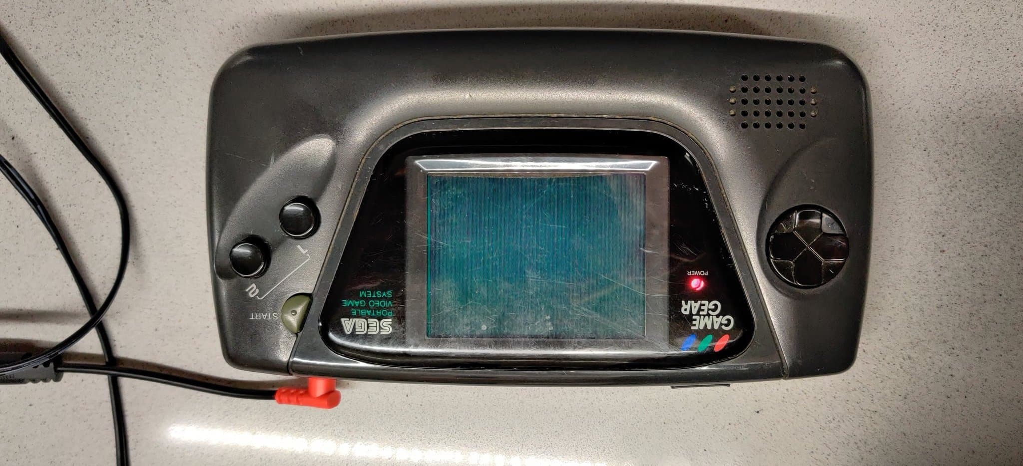 The second Sega Game Gear powered on, its screen black with no game picture but showing rows of stacked vertical lines, likely an artifact of the screen technology; this is the unit that boots and plays the Columns audio while showing no image.