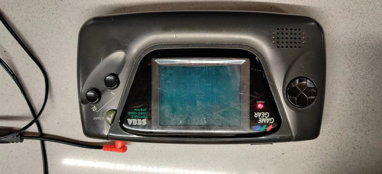 The second Sega Game Gear powered on, its screen black with no game picture but showing rows of stacked vertical lines, likely an artifact of the screen technology; this is the unit that boots and plays the Columns audio while showing no image.