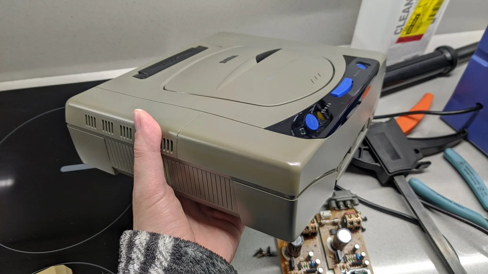 A hand holding the assembled Sega Saturn Model 1 console before retrobriting, the yellowing clearly visible on this side (the more yellowed of the two), with the recapped XR258 power board and hand tools on the worktop beside it.