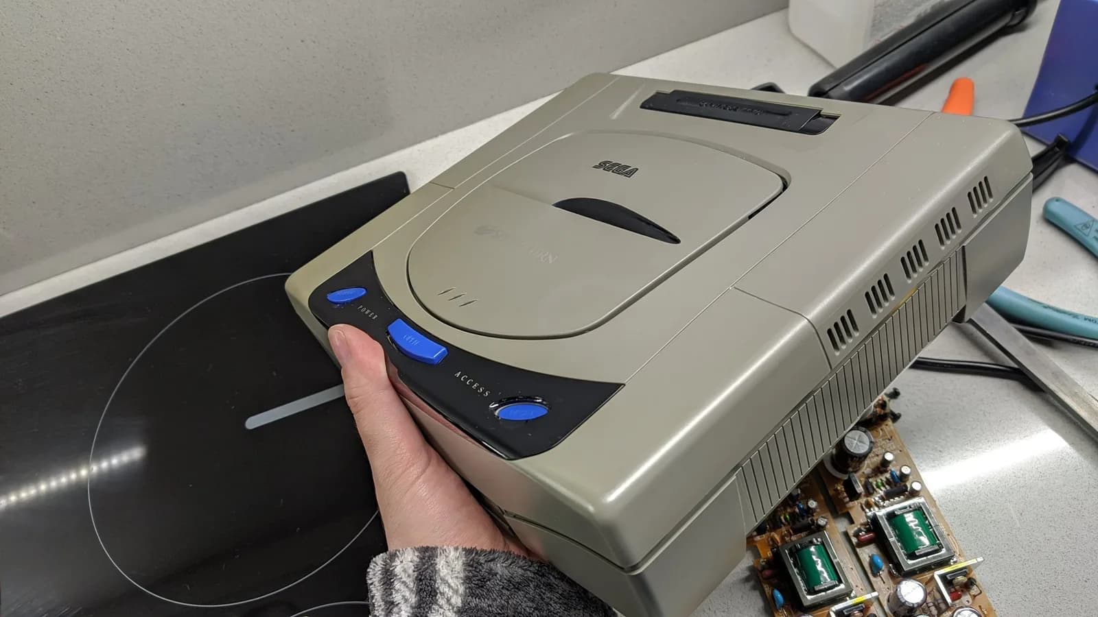The same Sega Saturn Model 1 console held from the other side before retrobriting, still yellowed but less so, with the recapped XR258 power board (two green transformers) and hand tools on the worktop beside it.