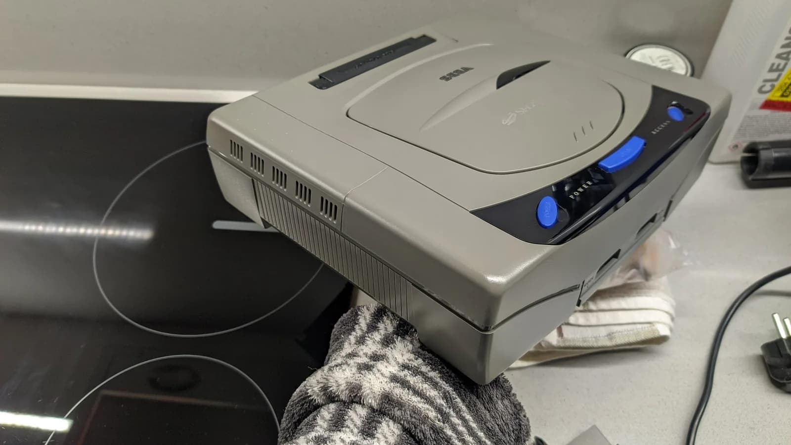 The Sega Saturn Model 1 console held after retrobriting, the side that had been the most yellowed now brought back to an even grey, over a black glass induction cooktop, with a bottle of isopropyl in the far background.