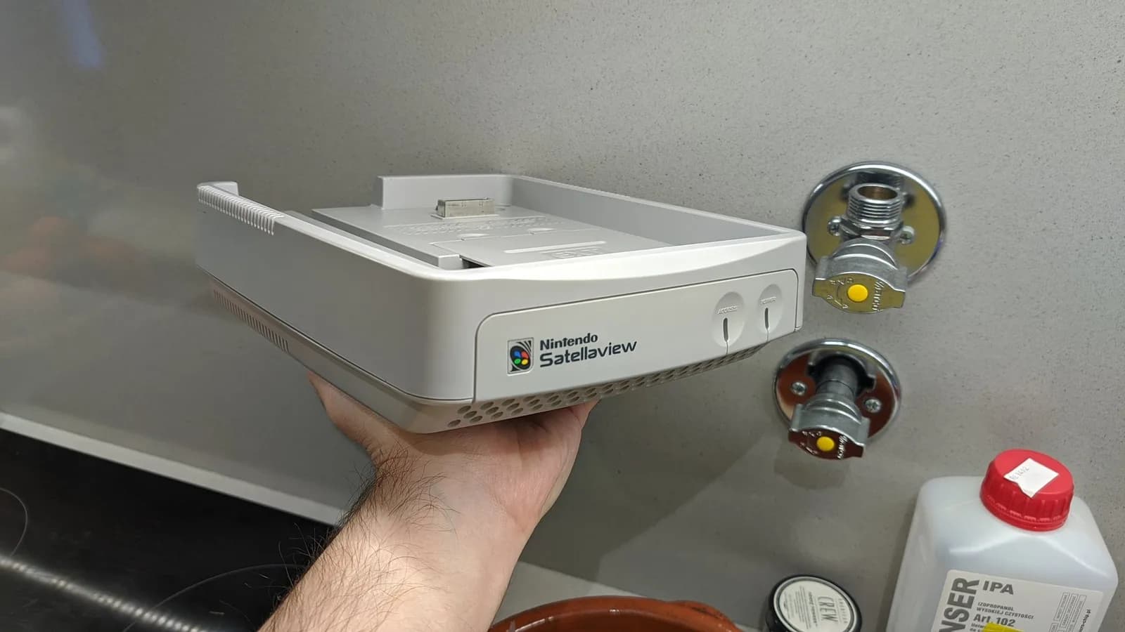 Nintendo SatellaView console held in hand after retrobriting, shell displaying improved whiteness; various electronic components visible nearby on workbench.