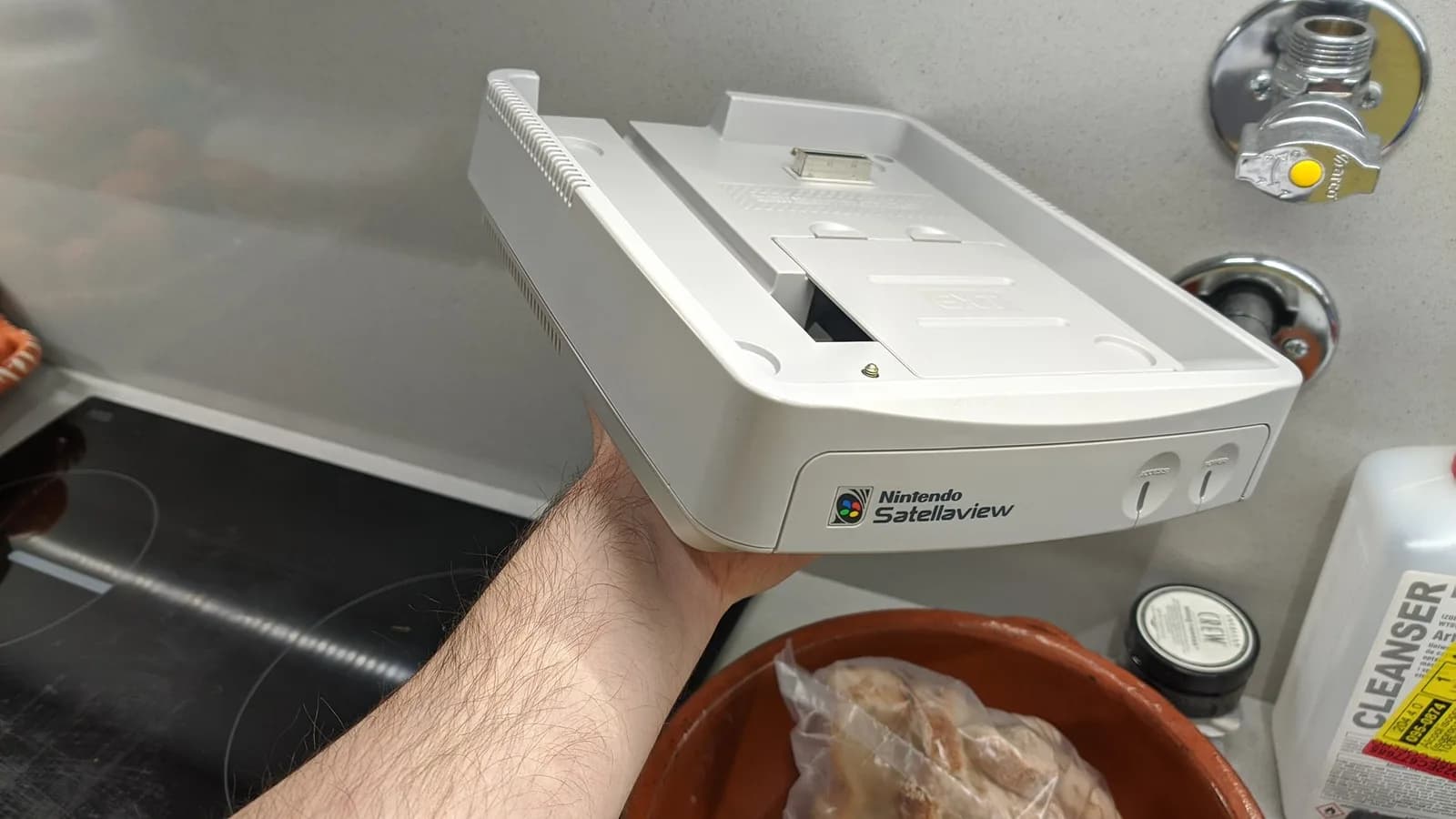 Hand holding white Nintendo SatellaView unit showing retrobriting results, shell partially disassembled beside work supplies on shelf.