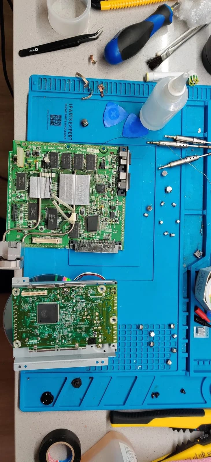 Sega Dreamcast VA0 motherboard (silkscreen KATANA MAIN VA0) and the optical-drive PCB on a blue silicone mat with all their electrolytic capacitors removed, ready for the new ones, the desoldered capacitors scattered around alongside tweezers, a screwdriver, a brush, a squeeze bottle and opening picks.