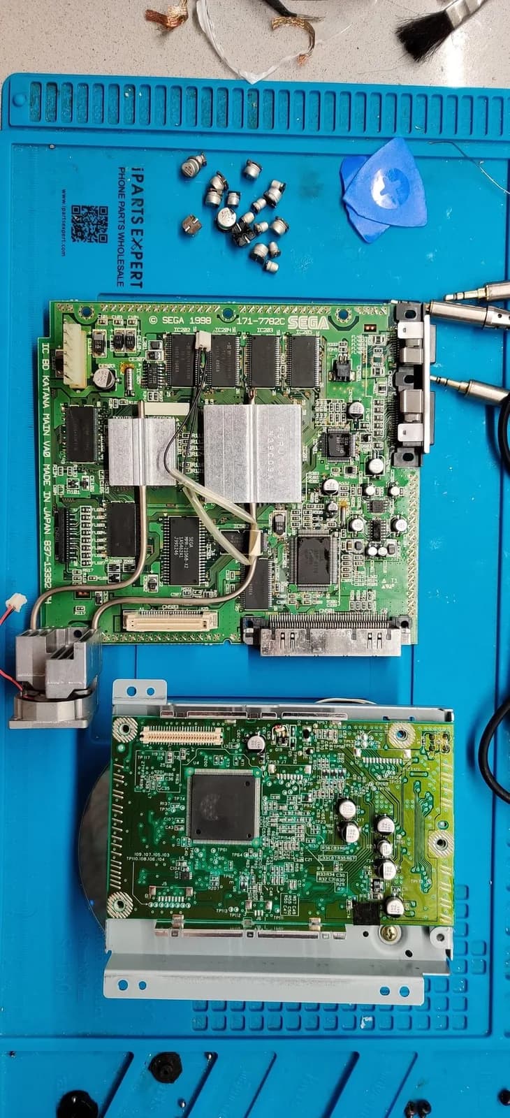 Sega Dreamcast VA0 motherboard (KATANA MAIN VA0) and the optical-drive PCB on a blue silicone mat, both now fully recapped with their new capacitors fitted, a blue opening pick and soldering iron tips to the side.
