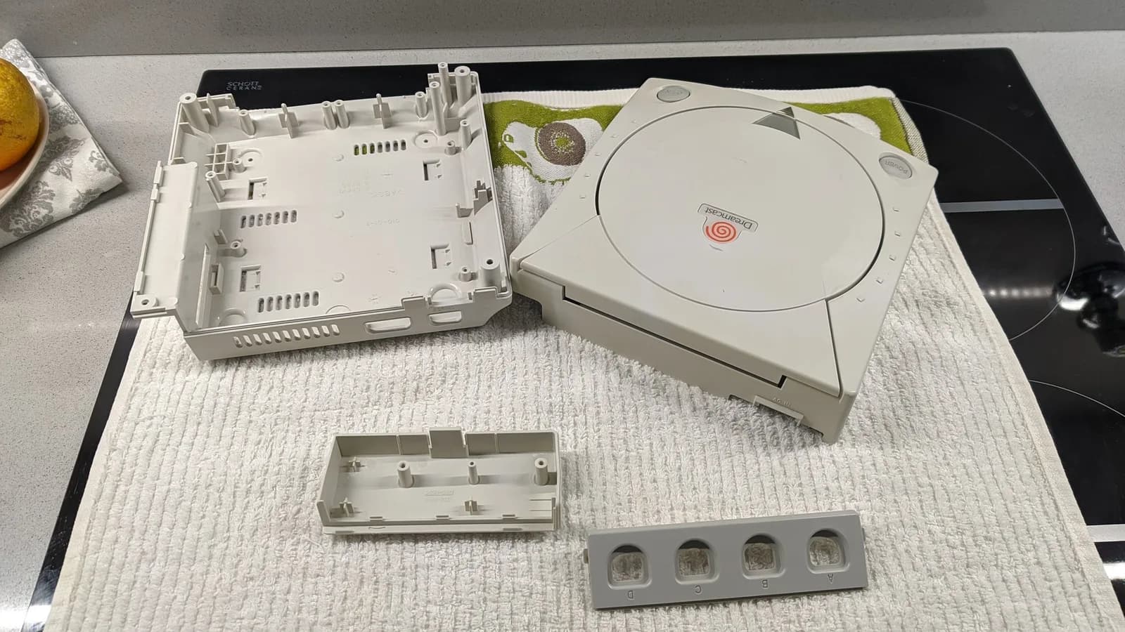 Sega Dreamcast VA0 shell parts laid out on a towel before retrobriting: the top case half (interior up), the main white shell body, a small internal bracket and the controller-port bezel labelled A B C D.