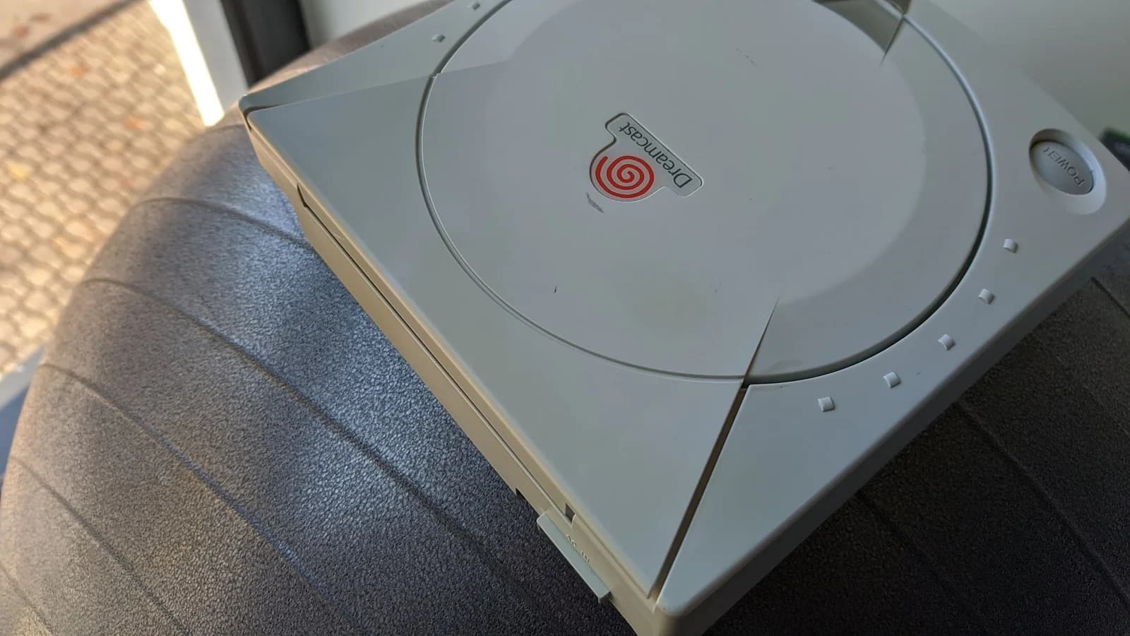 The Sega Dreamcast VA0 console before retrobriting, lid closed, photographed top-down on a dark surface near a window, its white shell yellowed with age.
