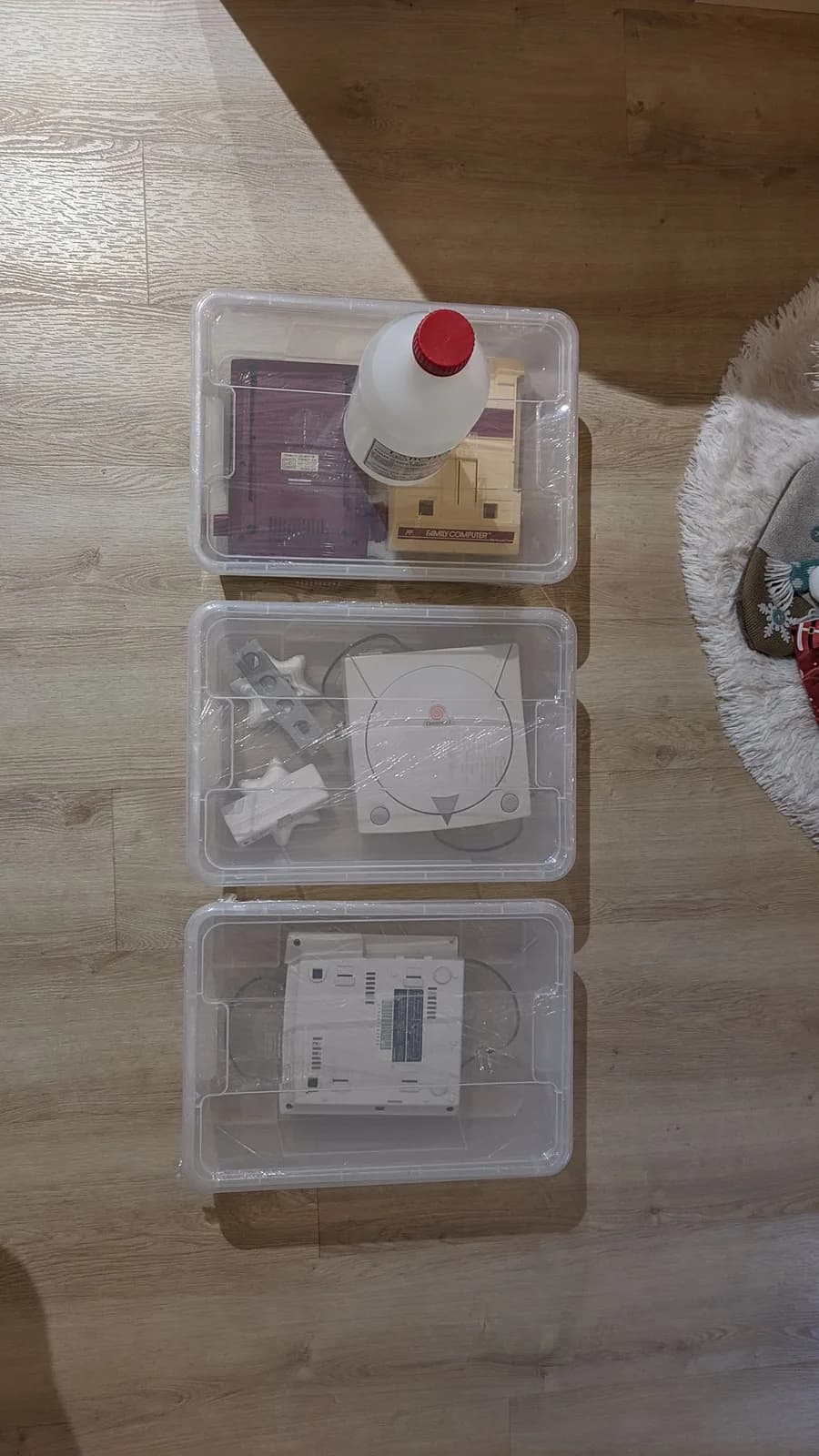 Three translucent bins on a wood floor staged for vapour-method retrobriting: two hold the disassembled Sega Dreamcast VA0 shell halves, and the third holds a Famicom shell with a red-capped bottle of hydrogen peroxide on top.