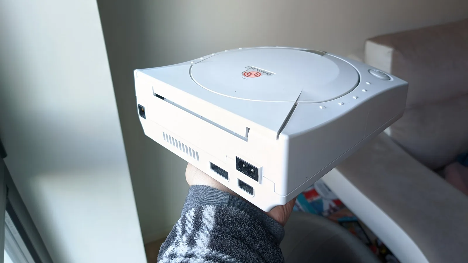 A hand holds up the rear of the white Sega Dreamcast VA0, freshly retrobrited, showing the AC power inlet, the rear AV and serial ports and the side cooling vents.