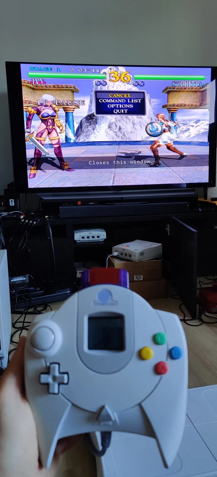 A hand holds a white Sega Dreamcast controller in front of a TV running Soul Calibur (Ivy versus Sophitia, Stage 1) with the pause menu open; the Dreamcast being played sits on cardboard boxes on the floor, with a second Dreamcast on a shelf.