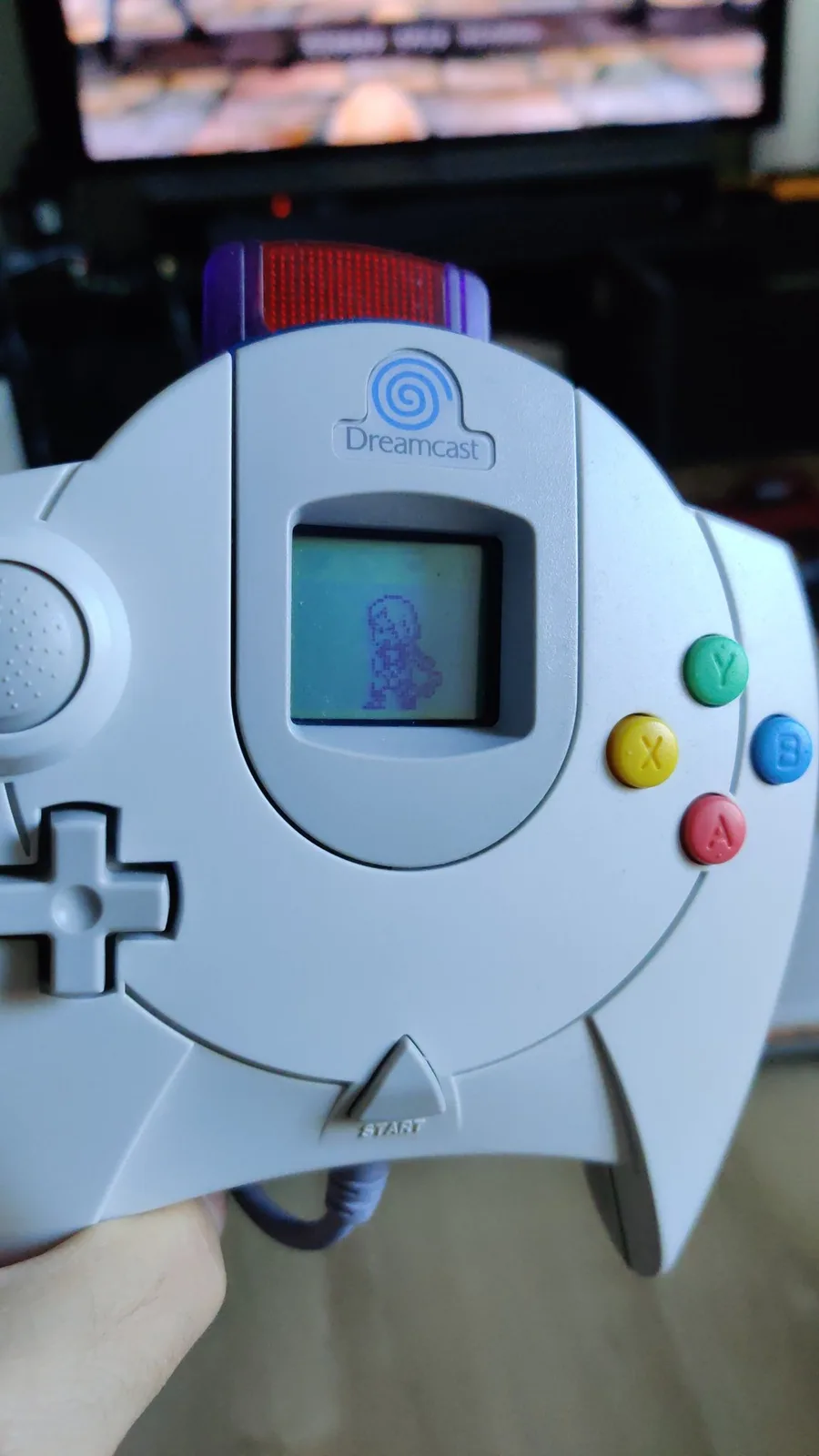 Close-up of a white Sega Dreamcast controller, its inserted VMU screen lit and showing a small purple character, with the green, yellow, red and blue face buttons and the d-pad; a Soul Calibur match plays on the blurred TV behind.