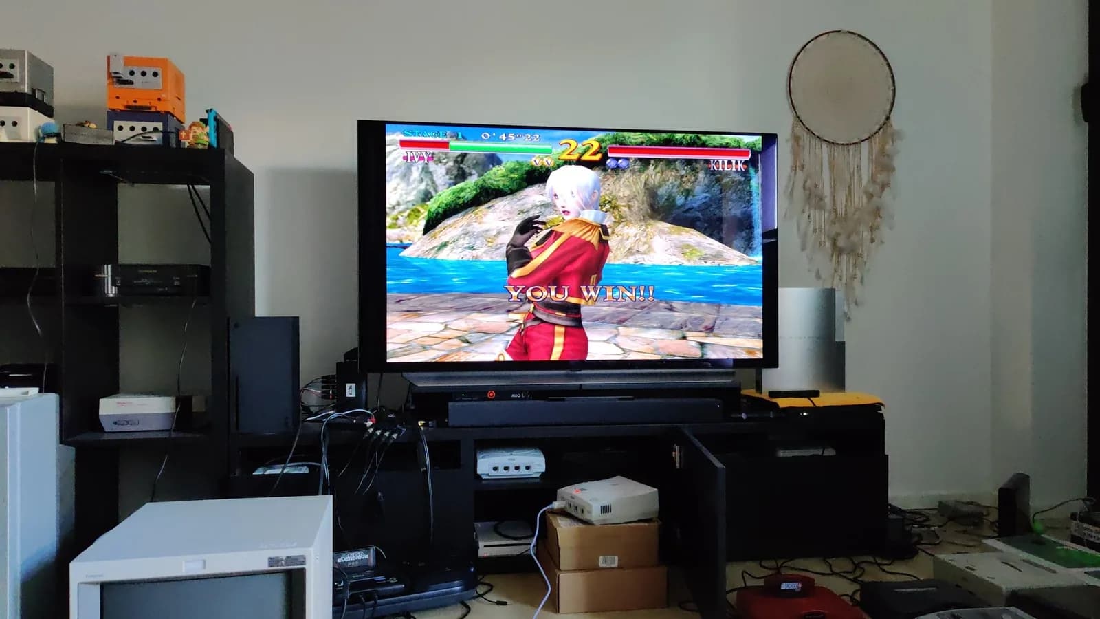 A flatscreen TV shows the Soul Calibur 'YOU WIN!!' victory screen (Ivy versus Kilik); a white Sega Dreamcast VA0 sits on the shelf below among other retro consoles.