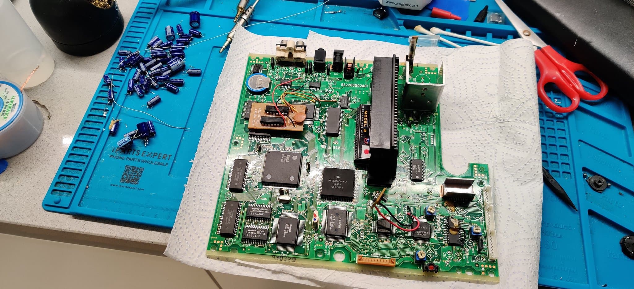 Sega Mega-CD 2 mainboard seen from the top side, fully decapped with all its electrolytic capacitors removed and a new region-free BIOS fitted, on the blue workmat.