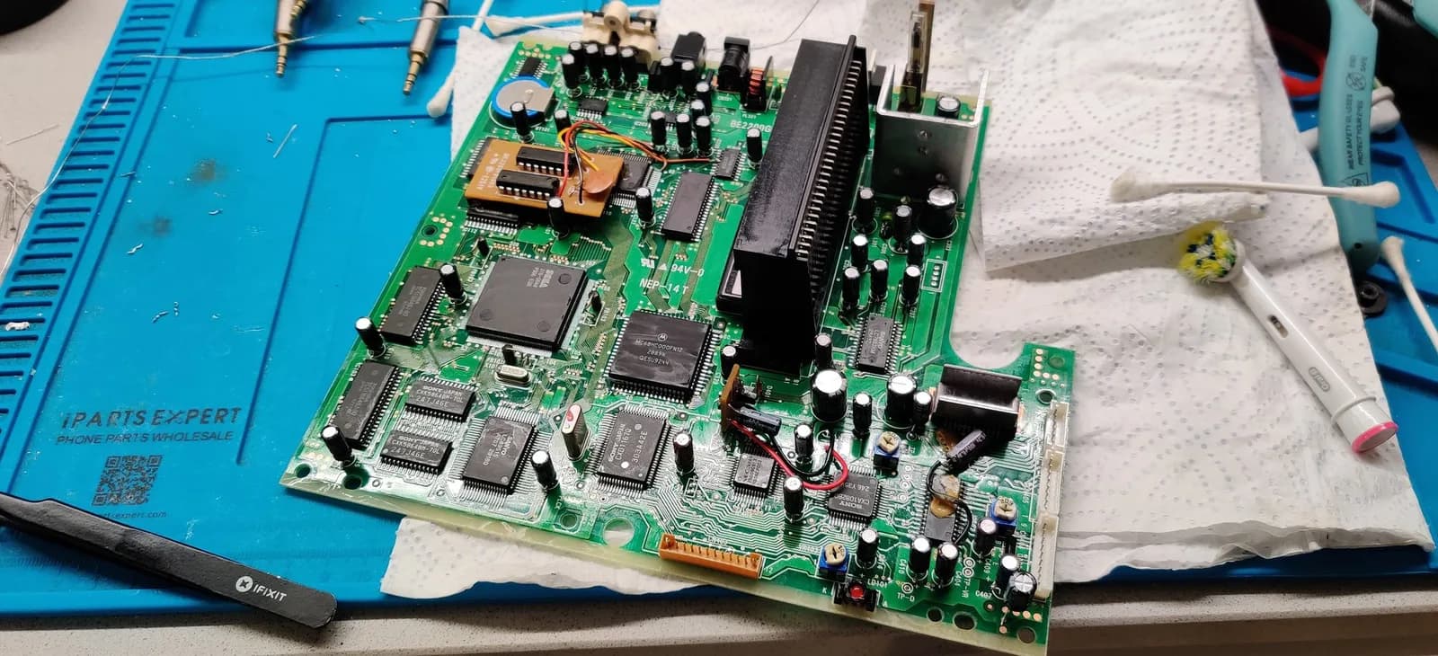 Sega Mega-CD 2 mainboard during recapping repair, showing replaced capacitors, IC chips, and heatsink with cleaning supplies on work mat.