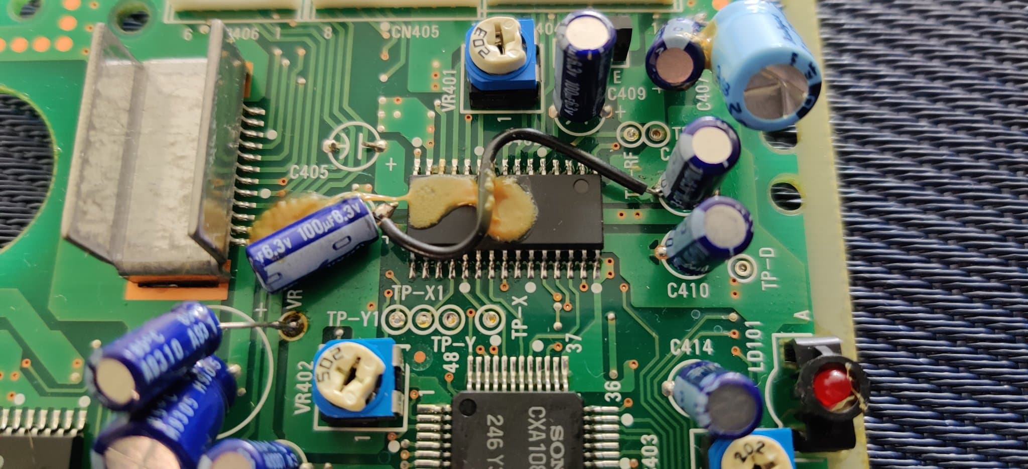 Close-up of the Sega Mega-CD 2 board showing the factory bodge wires and various ICs, with the electrolytic capacitors held down by factory glue or resin (not damaged), including a blue capacitor marked 100uF.