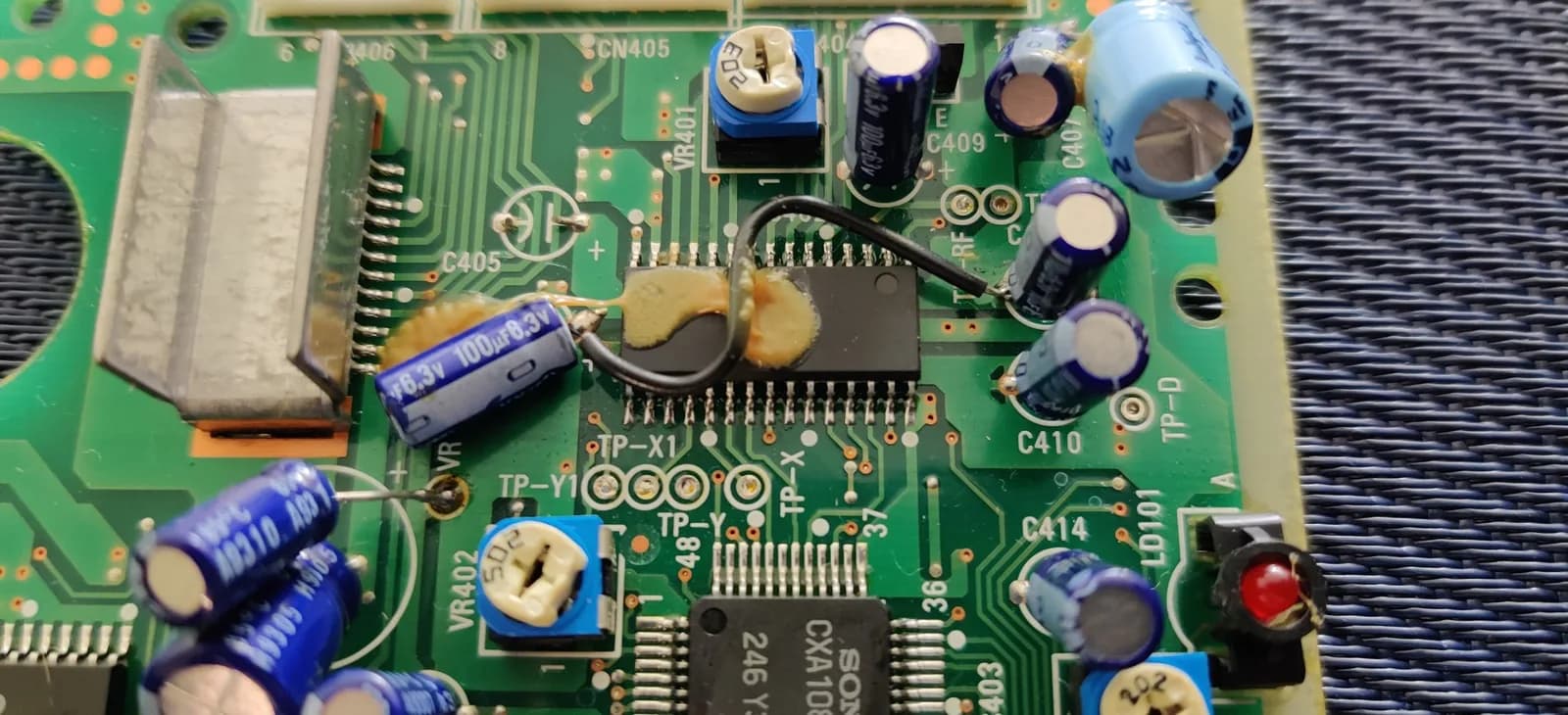 Close-up of the Sega Mega-CD 2 board showing the factory bodge wires and various ICs, with the electrolytic capacitors held down by factory glue or resin (not damaged), including a blue capacitor marked 100uF.