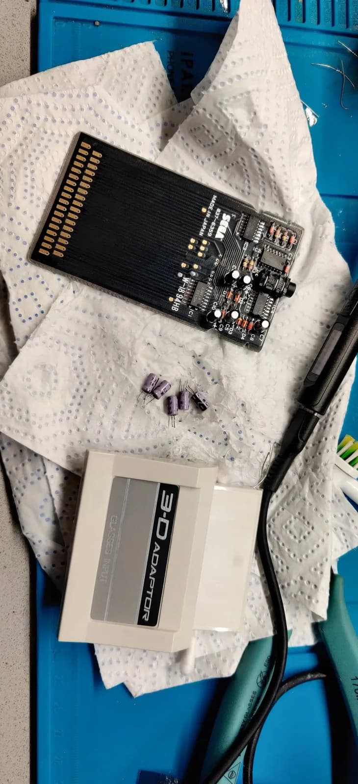 The Sega 3-D Glasses adapter PCB (silkscreen SEGA, 837-6395, Made in Japan) on a blue silicone mat during its recap, five removed purple electrolytic capacitors on the paper towel beside it, and the adapter's own white shell (labelled 3-D ADAPTOR / GLASSES INPUT) behind.