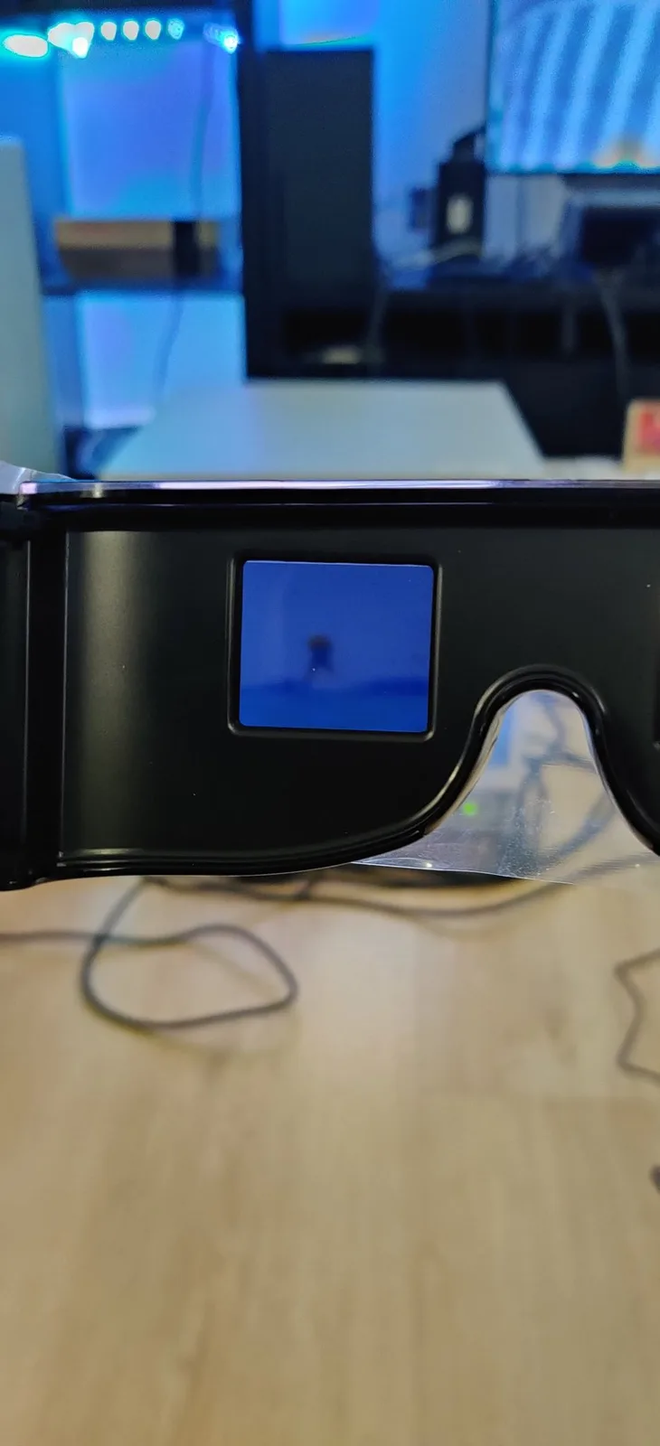 The black Sega active-shutter 3-D glasses (the eyewear) held up by a hand out of frame, the game showing in blue through the left LCD-shutter lens, an LED-lit shelf of consoles glowing behind.