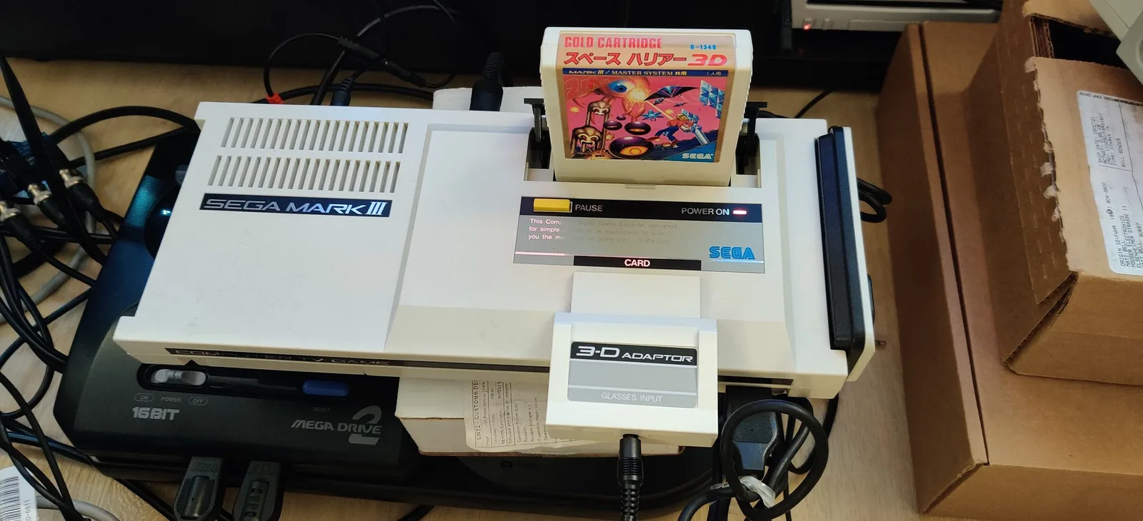 A beige Sega Mark III (POWER ON lit) with the white 3-D ADAPTOR / GLASSES INPUT card in its card slot and the Space Harrier 3D gold cartridge in the cartridge slot, stacked on a black Sega Mega Drive 2 and Mega-CD Model 2 combo, shipping boxes to the right.