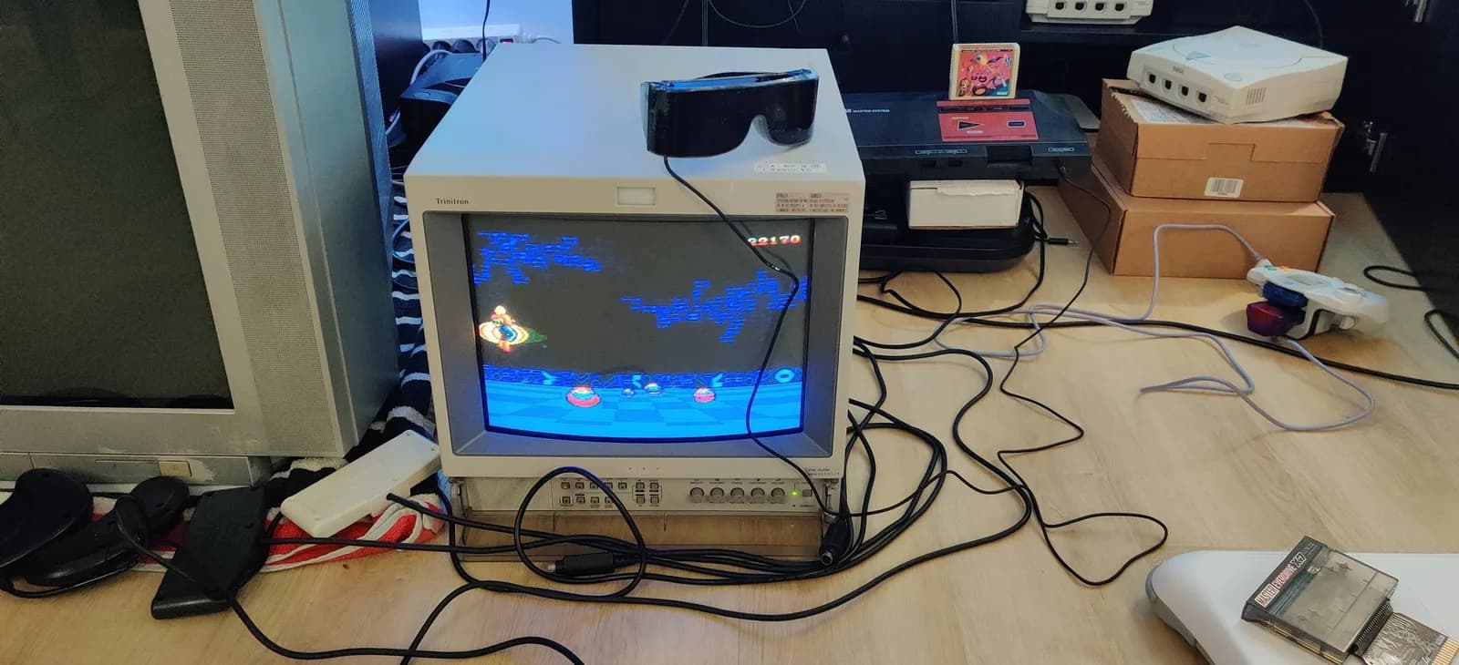 A Sony PVM-14L2 broadcast monitor running Space Harrier 3D (checkerboard ground, ring enemies, score 22170) with the black Sega shutter 3-D glasses resting on top of the monitor, and a Sega Master System (the Japanese first-revision MK-2000) with the Space Harrier 3D cartridge in its top slot, a Dreamcast and boxes around it.