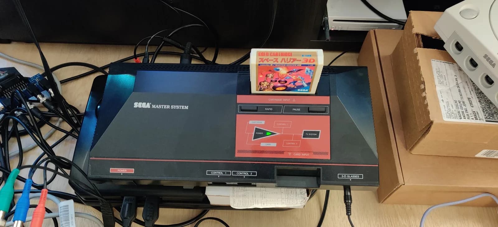 A Sega Master System (the Japanese first-revision MK-2000) with a built-in 3-D GLASSES port on the front and the Space Harrier 3D gold cartridge in the slot, cables to the left and a Dreamcast plus boxes to the right.