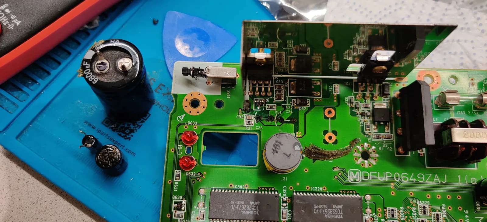 Panasonic FZ-1 motherboard showing power supply section with damaged traces, ceramic capacitor, and electrolytic capacitor requiring repair work.