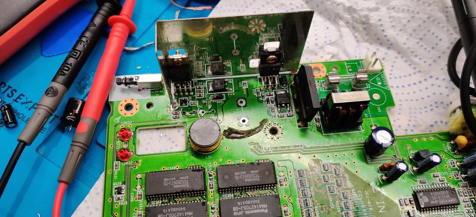 Panasonic FZ-1 motherboard with multimeter probes, solder wick, and desoldering tools during PSU trace repair and recapping restoration work.