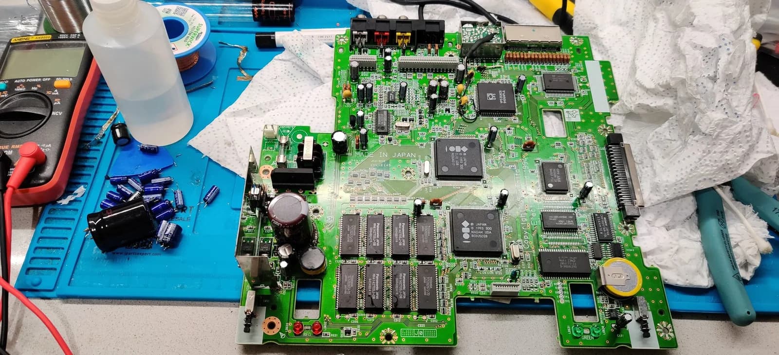 Panasonic FZ-1 mainboard undergoing PSU repair with multimeter, flux, solder braid, recapped board showing trace damage and bridged connections.