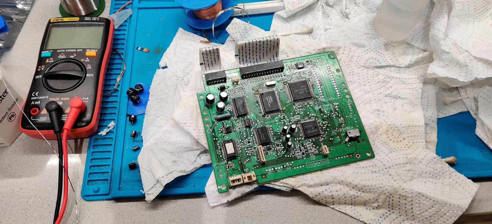 Panasonic FZ-1 optical drive PCB recapping: green circuit board with replaced capacitors, multimeter probing components, heat sinks and ICs visible on blue work mat.
