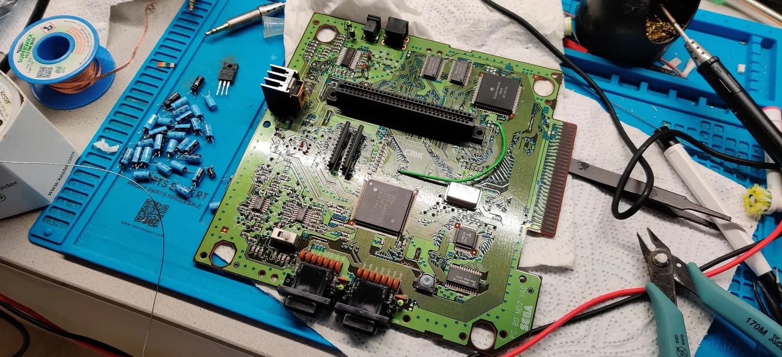 Sega Mega Drive II main board undergoing refurbishment: capacitors removed, triple bypass mod components visible, soldering iron and tools on blue mat workstation.