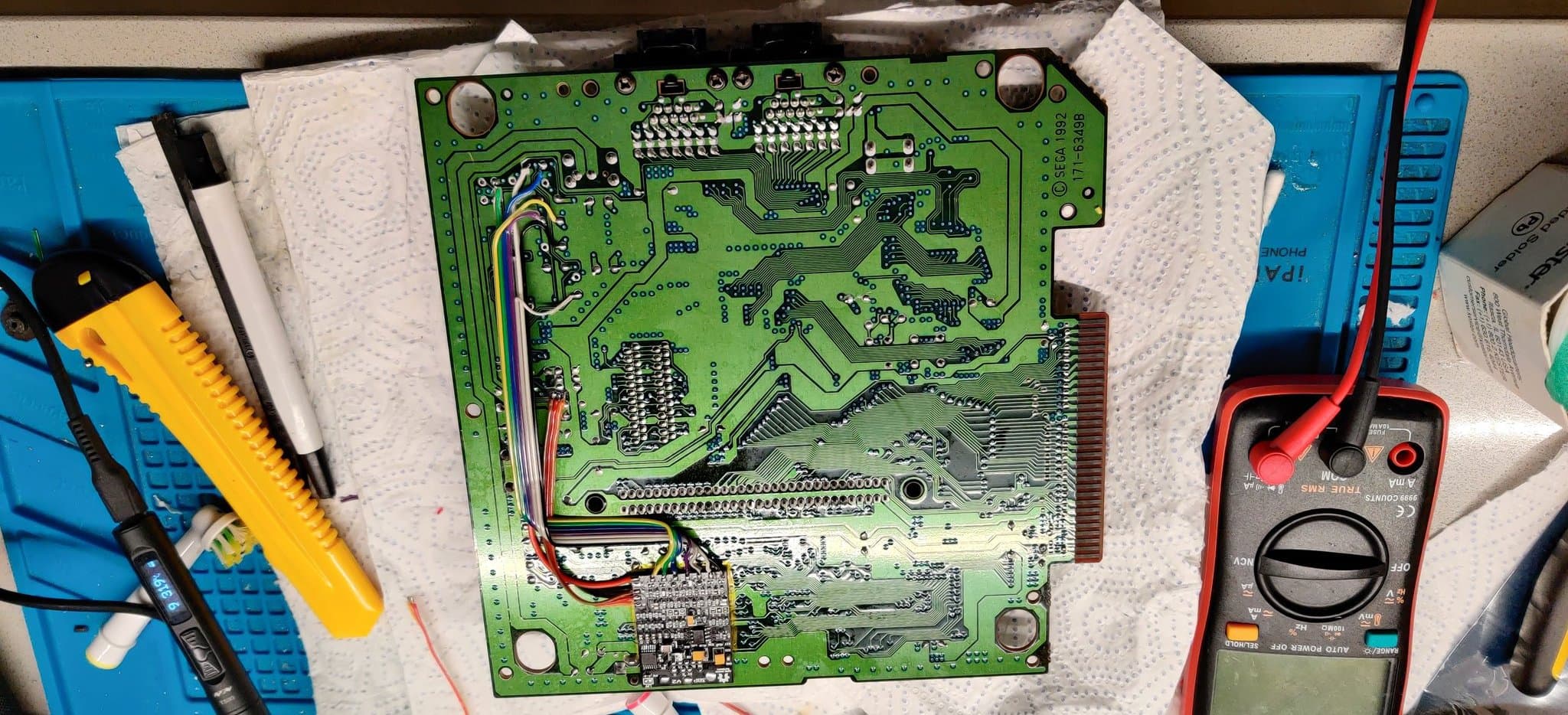 Sega Mega Drive II mainboard with capacitor replacement work in progress, multimeter and soldering iron visible, triple bypass mod installation underway.