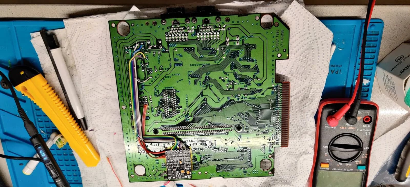 Sega Mega Drive II mainboard with capacitor replacement work in progress, multimeter and soldering iron visible, triple bypass mod installation underway.