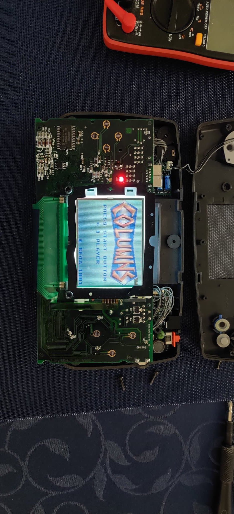 Sega Game Gear PCB with replaced capacitors and reflowed solder joints; LCD displays Sega logo with multimeter and soldering iron visible above.