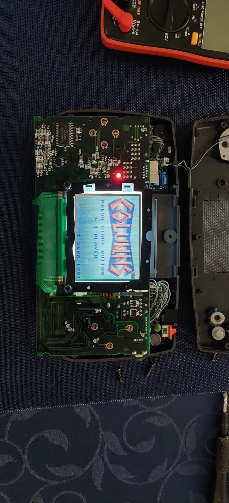 Sega Game Gear PCB with replaced capacitors and reflowed solder joints; LCD displays Sega logo with multimeter and soldering iron visible above.