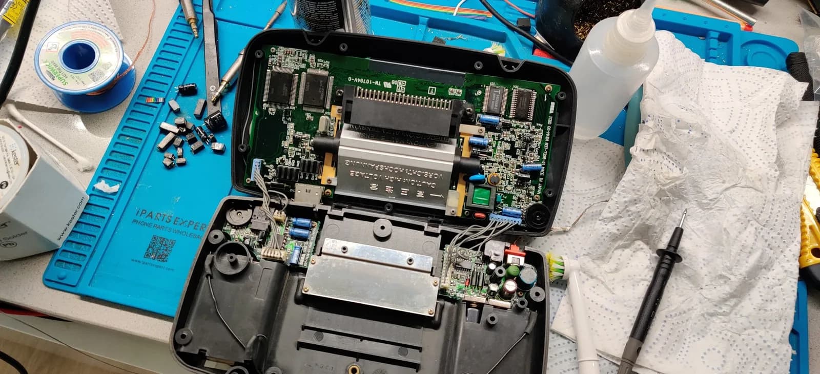 Disassembled Sega Game Gear motherboard with exposed capacitors, solder joints, and power board during restoration work on blue mat.