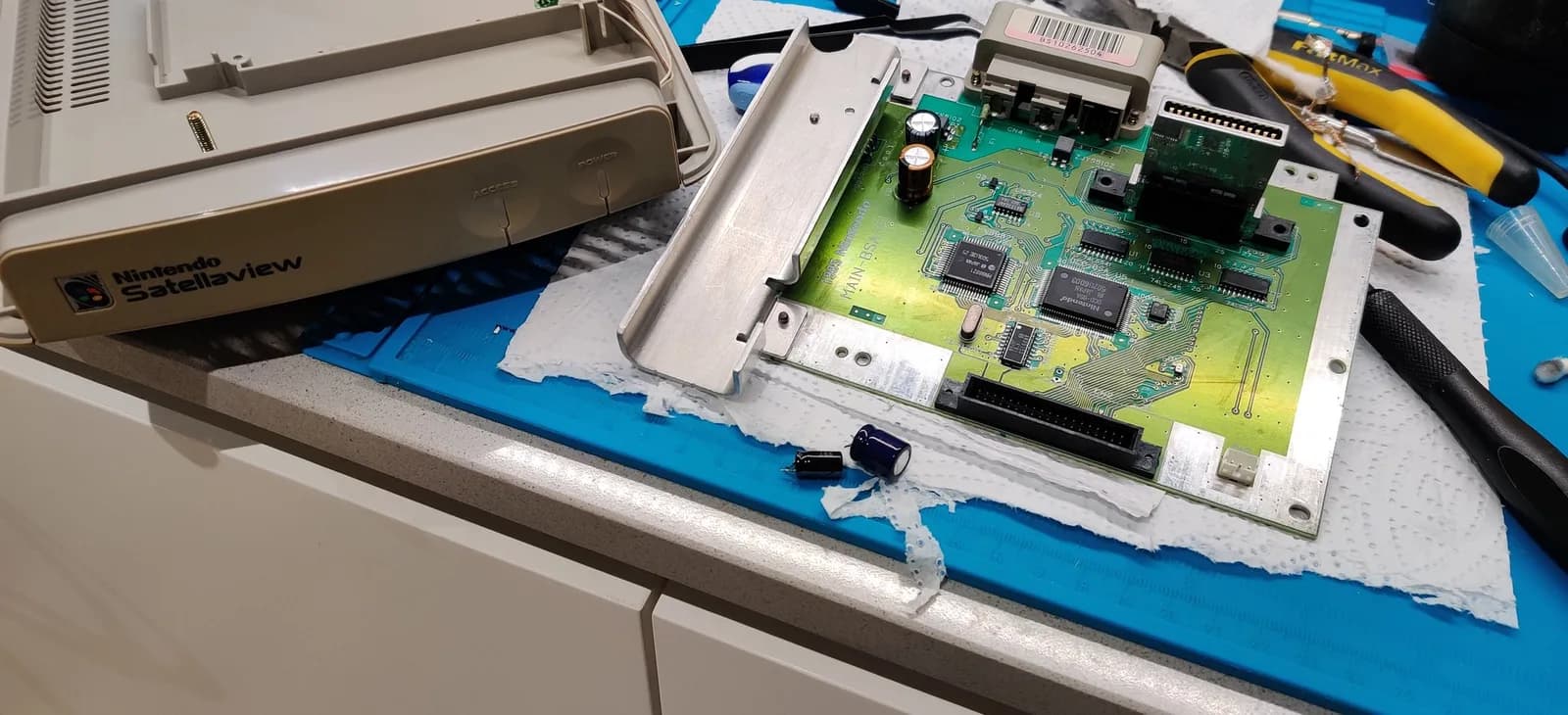 Nintendo Satellaview recap: the removed top shell half (Nintendo Satellaview logo) beside the green MAIN-BSX main board and the removed old electrolytic capacitors on a blue work mat.