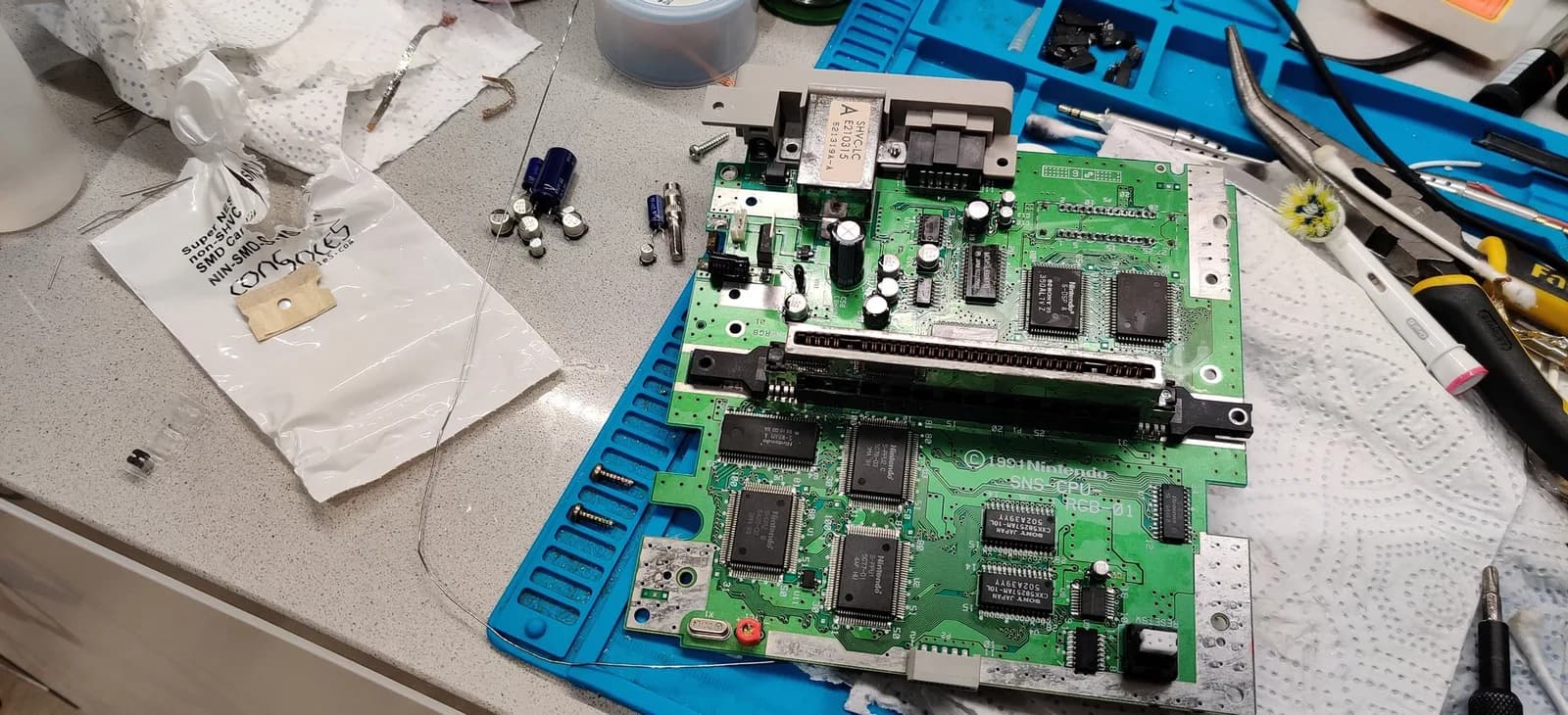 The recapped Super Famicom main board (metal SHVC-LC module) on a blue work mat, the removed old capacitors and a Console5 cap-kit bag on the grey countertop to its left.