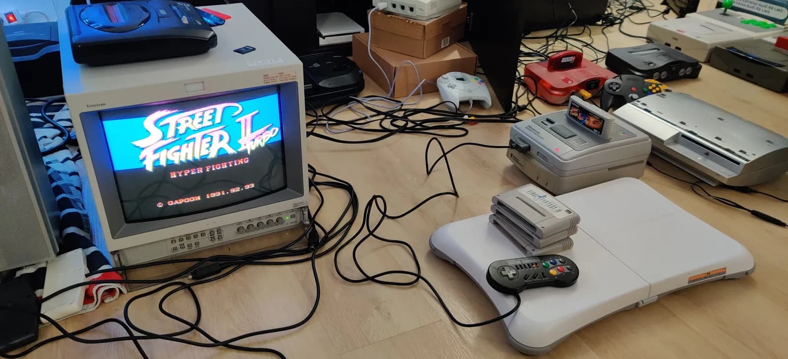 Repaired Nintendo Super Famicom (on a Nintendo Satellaview base, cartridge inserted) running Street Fighter II Turbo: Hyper Fighting on the Sony PVM-14L2; a Fighting Commander pad, stacked Super Famicom cartridges, and other consoles and controllers on the wood floor around it.
