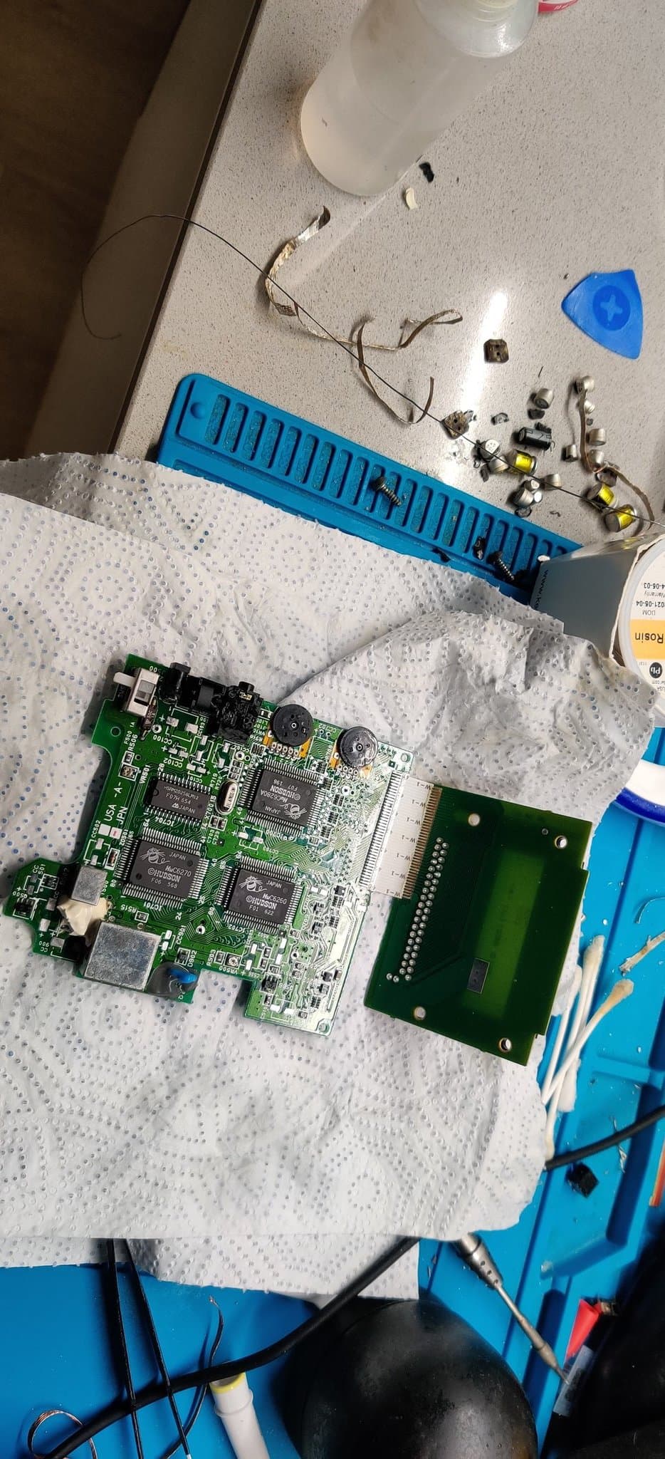 PC Engine GT boards with all capacitors removed by gently twisting the cans loose (A3rgan's method) and the pads cleaned with flux and braid without losing one, on the blue silicone mat beside the desoldering tools.
