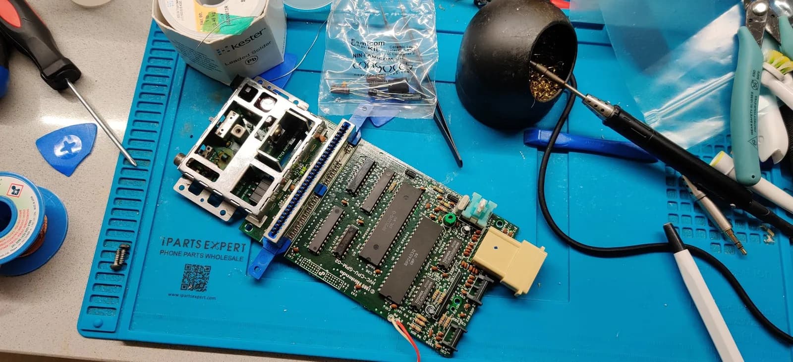 Famicom motherboard on work mat with capacitors, composite mod board, soldering iron, multimeter, and replacement fuse during repair and testing.