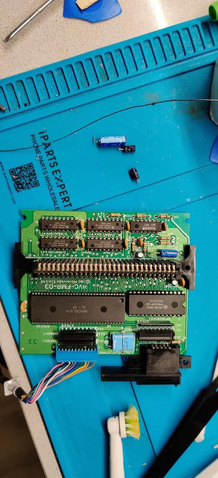 Famicom Disk System RAM adapter board (silkscreen HVC-FMR-03), component side up on a blue work mat with its through-hole chips and card-edge connector, and three old electrolytic capacitors that were desoldered lying on the mat just above it.