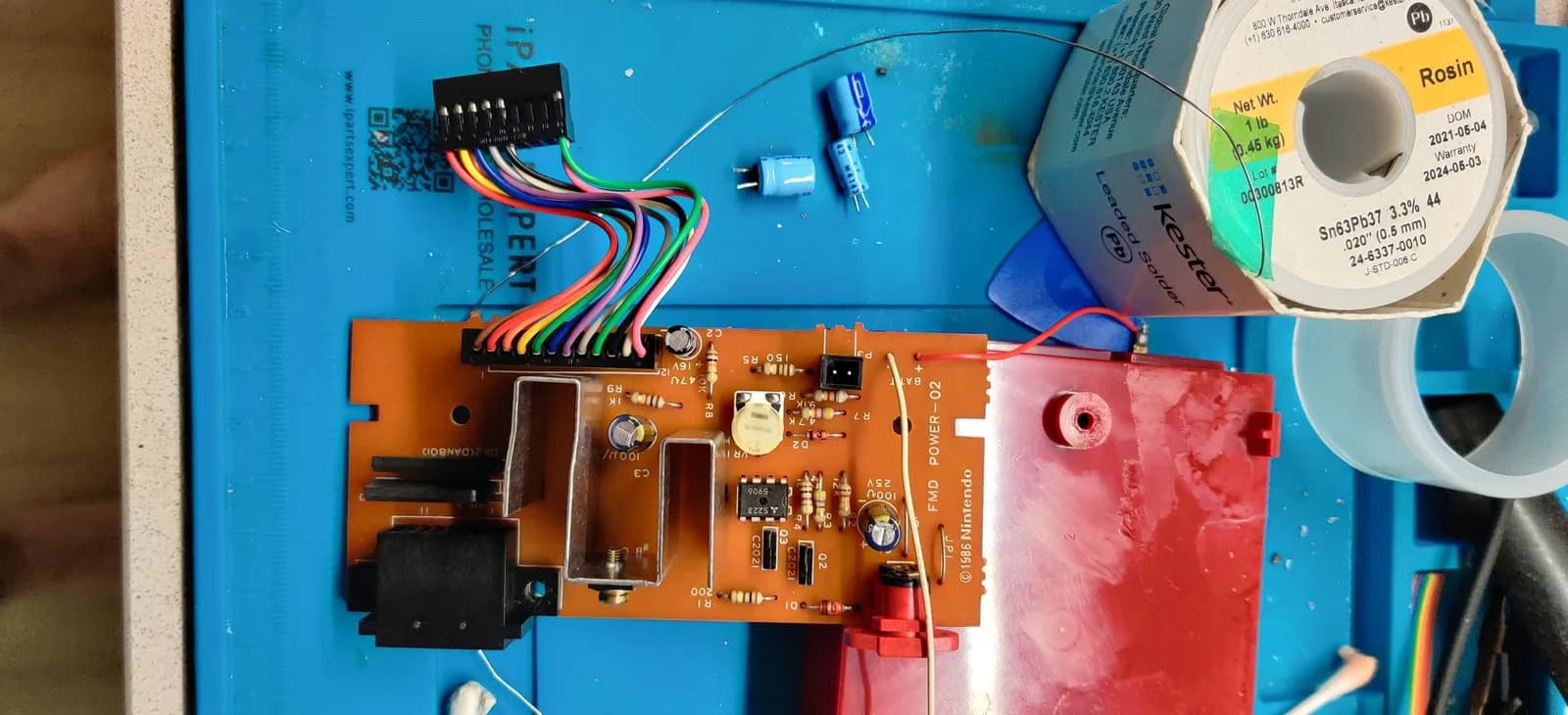 Famicom Disk System power board (silkscreen FMD POWER-02) being recapped: an orange phenolic PCB with a multicolored wiring harness on a black connector, the red plastic housing of the disk drive unit beside it at right, a Kester solder spool at top right, and the removed old blue electrolytic capacitors on the blue mat.