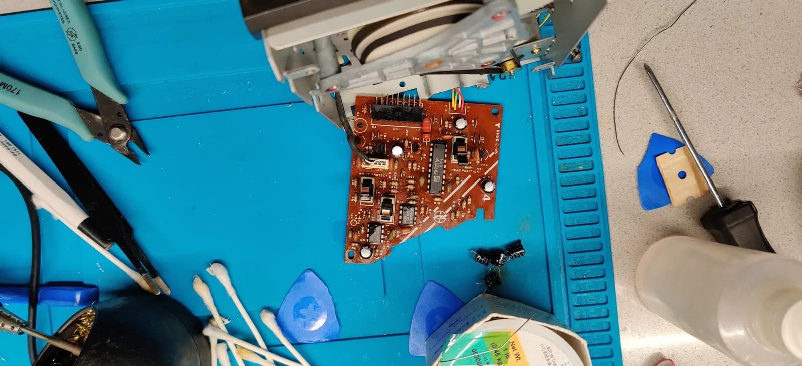 Famicom Disk System drive control board (silkscreen HEAD POSITION, WRITE PROTECT and MOTOR STOP) mid-recap on a blue mat, with several removed old electrolytic capacitors beside it; a soldering iron rests at the bottom left and a Kester solder spool at the bottom right, with flush cutters, tweezers and cotton swabs around it. The drive mechanism sits at the top edge, its newly fitted belt looped round the plastic flywheel.