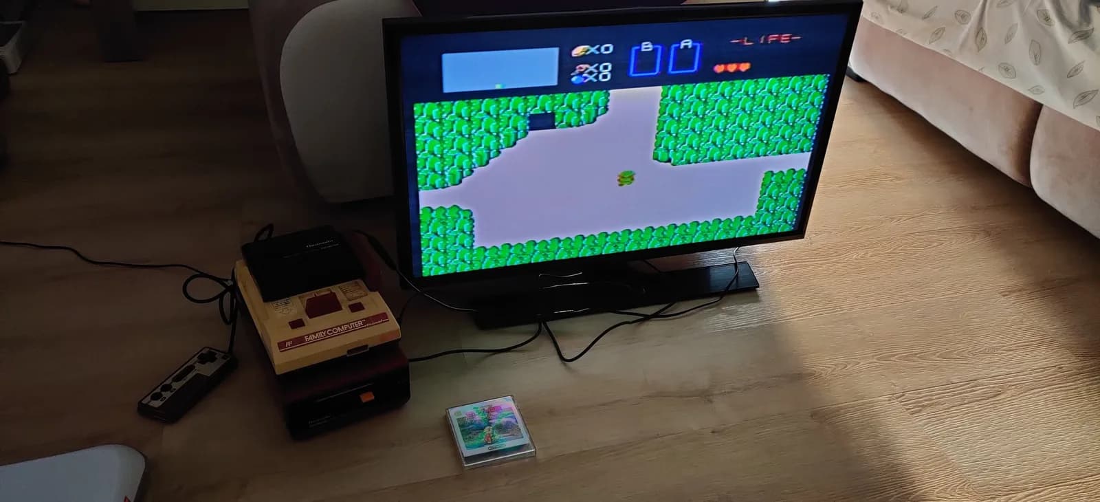 A flat-panel TV on a wooden floor shows The Legend of Zelda (Famicom Disk System) overworld gameplay after the refurbishment; at left the cream-and-red Famicom is stacked with its black Disk System RAM adapter and the disk drive next to a Famicom controller, and the empty case of the Zelda Disk System game sits on the floor in front, its disk loaded in the drive.