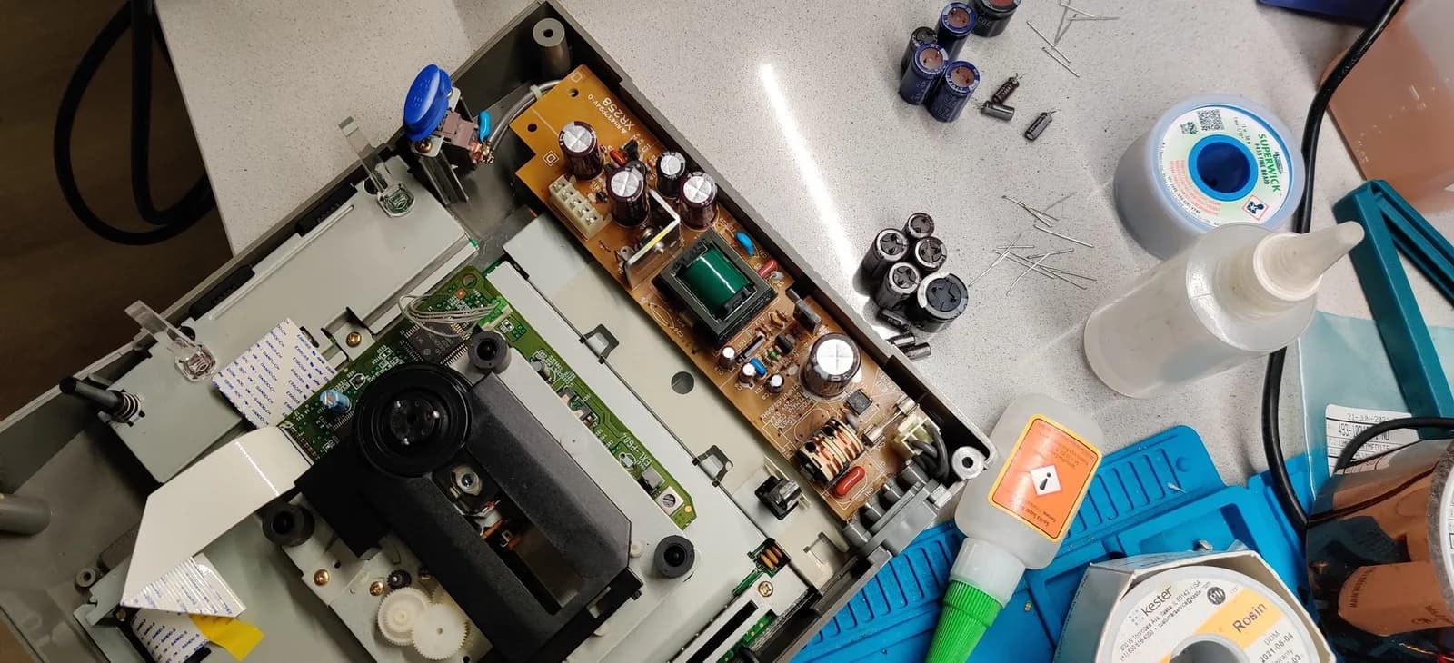 A disassembled Sega Saturn Model 1 on a worktop during the recap: the metal chassis with the optical-drive laser assembly and white gears on the left, the orange XR258 power board with a green transformer and electrolytic capacitors, the old pulled caps and snipped leads to the right, and a Superwick desoldering braid spool, flux and Kester solder on a blue silicone mat.