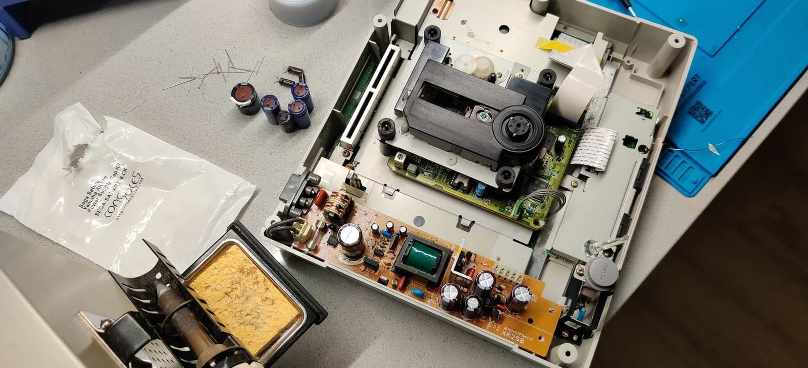 A disassembled Sega Saturn Model 2 on the bench, its orange XR258 power board already recapped (fresh capacitors fitted): the inductor coil, green transformer and new caps on the board, the optical-drive laser assembly and white gears above it, the old pulled caps displayed nearby, a soldering iron in its stand, and an open Console5 Sega Saturn Yamaha XR258 power supply cap kit bag to the left.