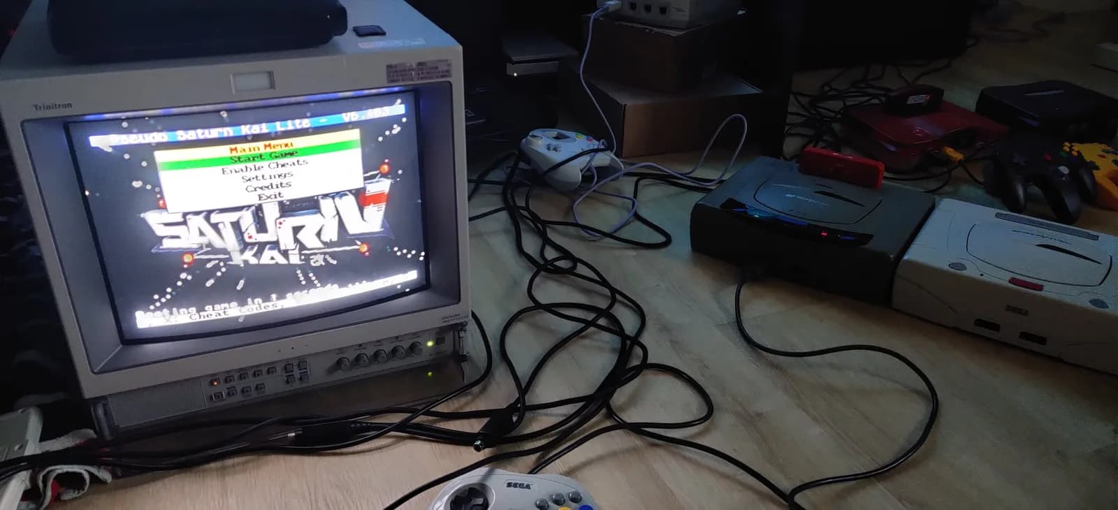 A grey Sega Saturn Model 1 (with a red Pseudo Saturn Kai cartridge in its slot) connected to a Sony PVM-14L2 broadcast monitor showing the Pseudo Saturn Kai Lite V6 homebrew boot menu (Main Menu, Start Game, Enable Cheats, Settings, Credits, Exit), a white Saturn controller and other consoles on the wood floor during the post-recap smoke test.